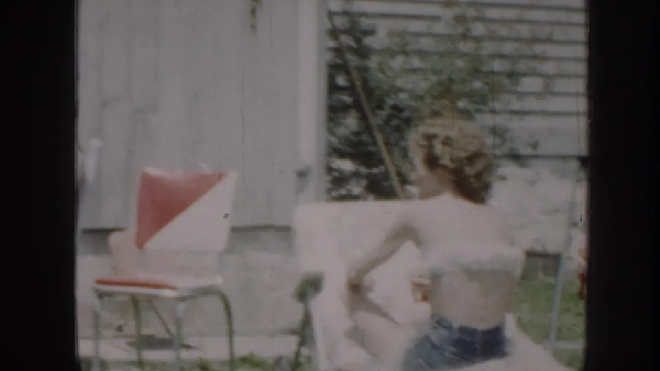 1958 Nebraska home-movie frame showing a woman reclining in a backyard lawn chair beside a red-and-white outdoor chair.