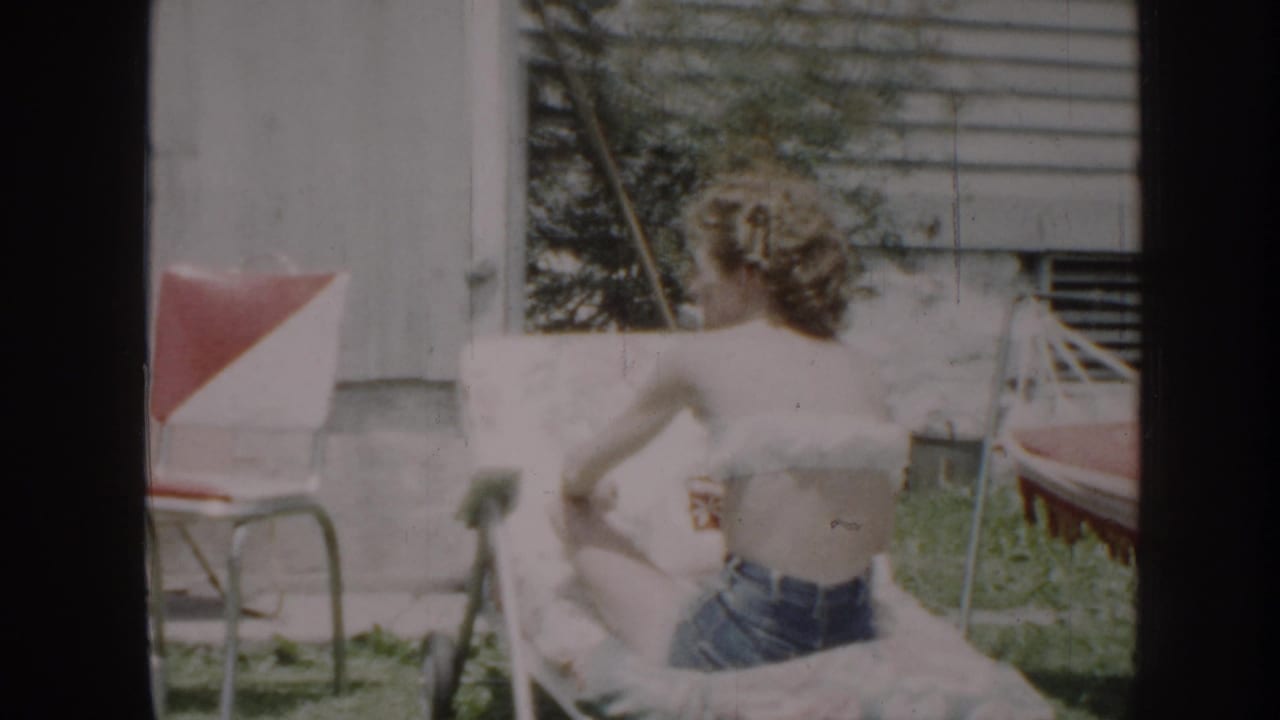 Woman in jeans reclines on a lawn chair beside outdoor furniture and house siding.