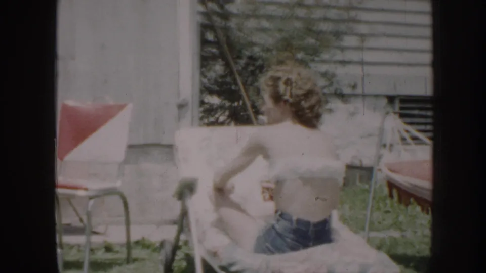 A woman reclines in the sun in a Nebraska backyard, with lawn furniture and house siding visible behind her.