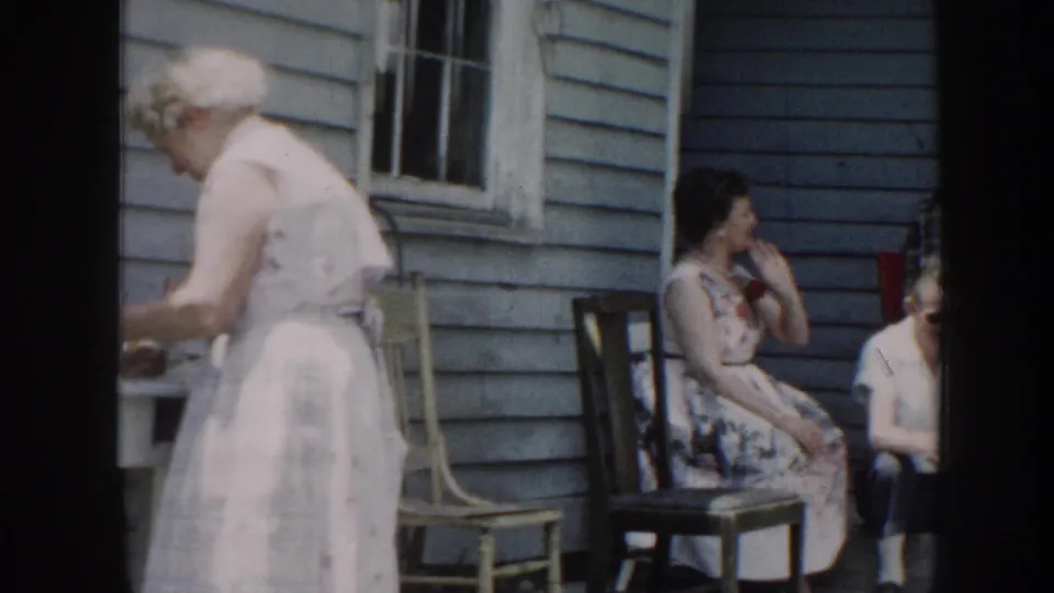Nebraska backyard gathering with a woman at a table and adults seated near a blue-sided house, filmed on 8mm in 1958.