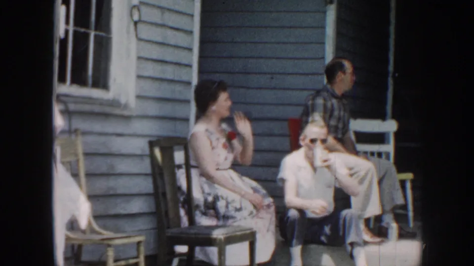 Adults gather outside a blue-sided Nebraska house, seated in casual backyard chairs during a 1958 home-movie scene.