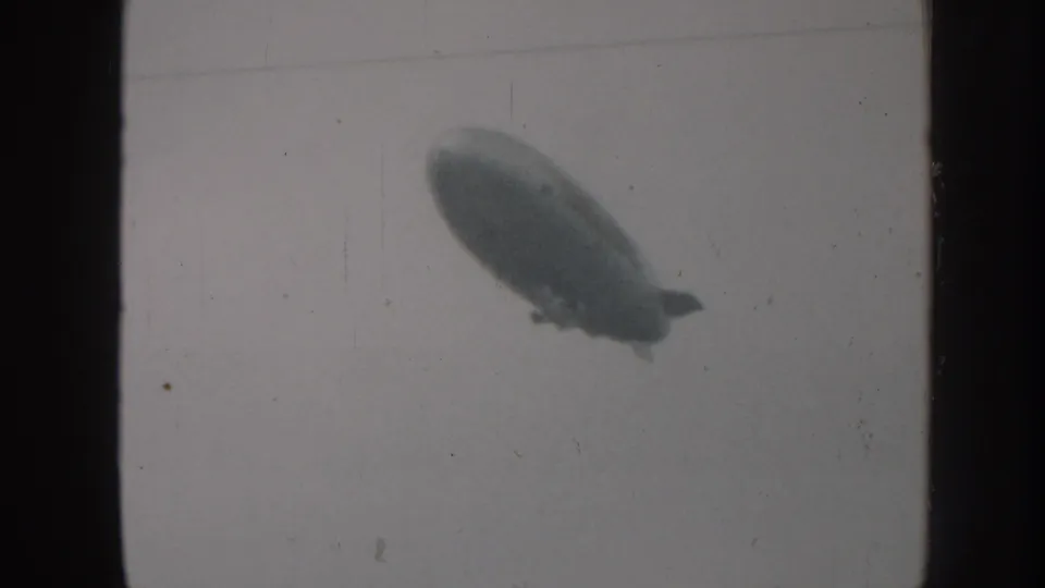 1958 Nebraska 8mm frame showing a blimp-like dirigible gliding through an overcast sky with visible film grain.
