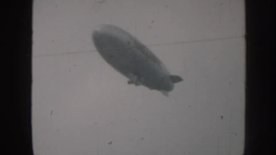 1958 Nebraska home-movie frame with the airship closer or larger in view, softened by 8mm grain and light scratches.
