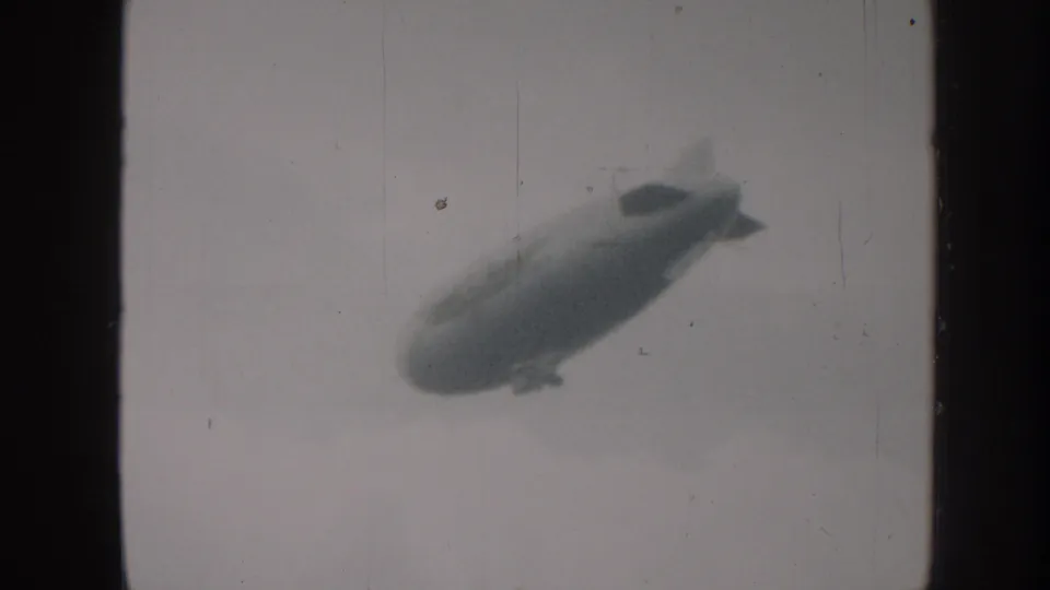1958 Nebraska 8mm frame of a large airship angled across an overcast sky, bordered by dark film edges.
