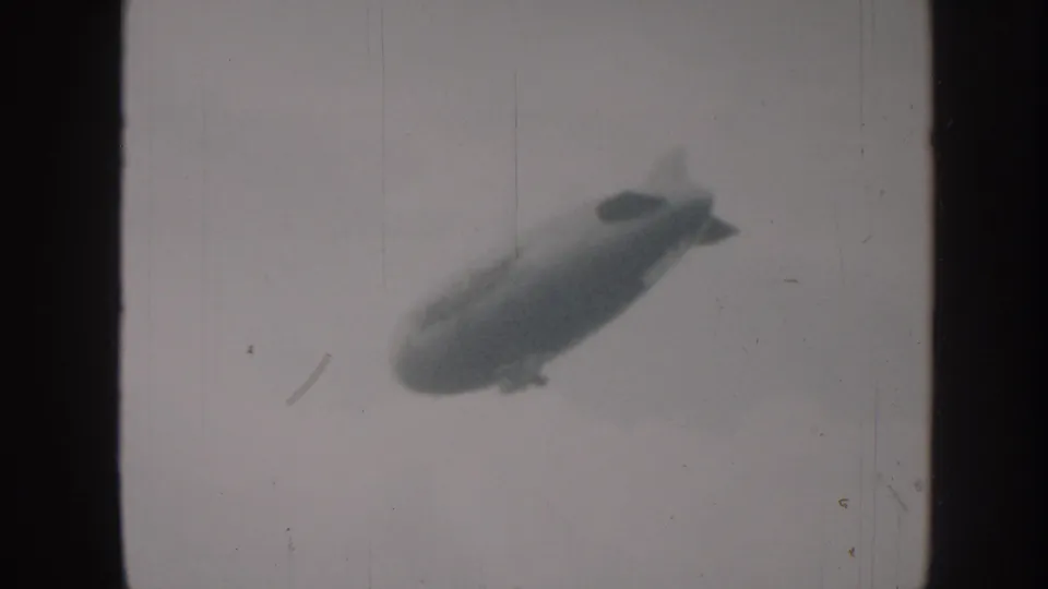 A large blimp appears as a soft silhouette against a cloudy Nebraska sky in this 1958 home-movie frame.
