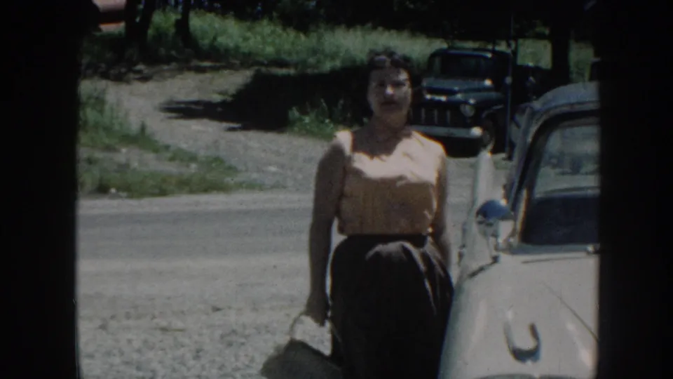 A woman stands beside a light-colored 1950s car on a roadside in Nebraska, with another vintage vehicle in the background.