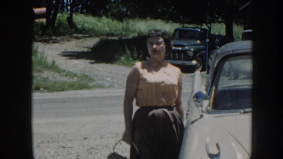 Adult woman in 1958 Nebraska footage stands near a white car, holding a small handbag with trees and a dark pickup behind her.