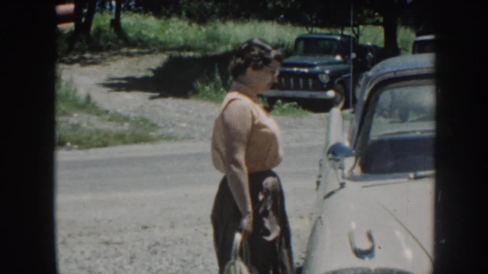 Adult woman turns in profile near a white car on a rural Nebraska road, with grass, trees, and a dark pickup in the background.