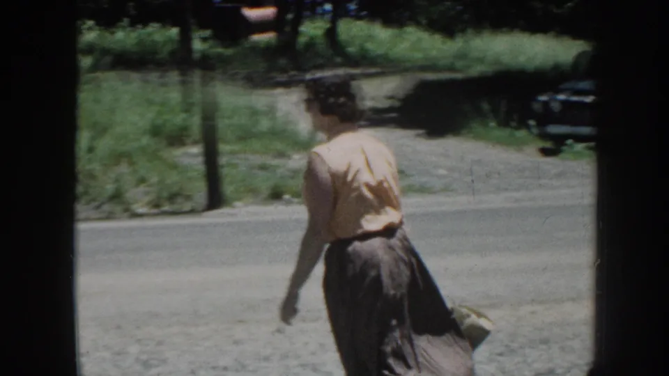 Adult woman walks away from the car in 1958 Nebraska home-movie footage, carrying a small handbag beside a rural road.