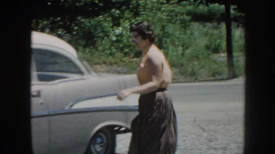 Adult woman passes the side of a white vintage car in a rural Nebraska setting, framed by grass and roadside foliage.