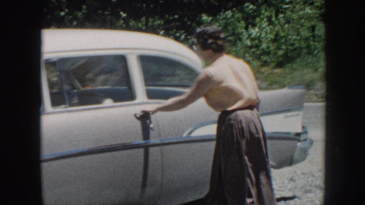 Woman in a pink sleeveless top opens the door of a gray sedan beside a gravel roadside.