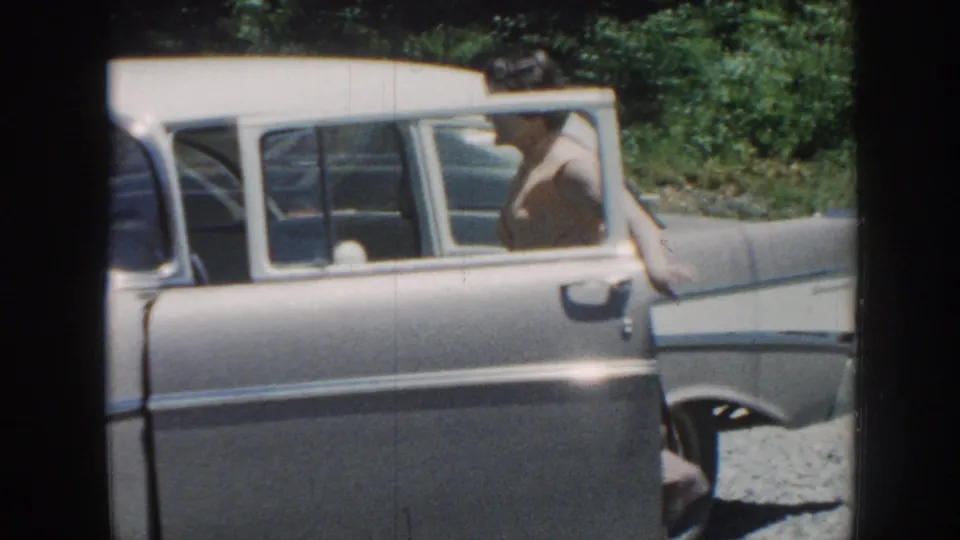 A woman appears to climb into a gray 1950s sedan with the door open during a sunny Nebraska home-movie scene.