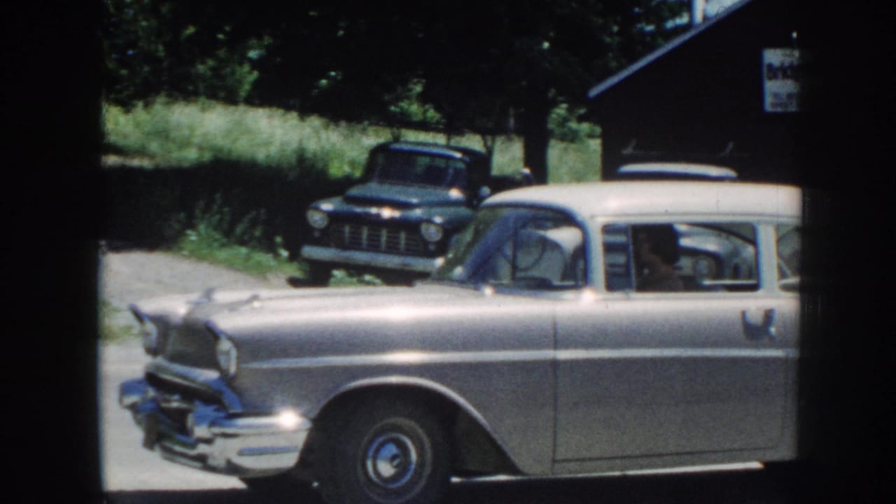 Silver 1950s sedan passes a dark pickup near trees and a wooden building.