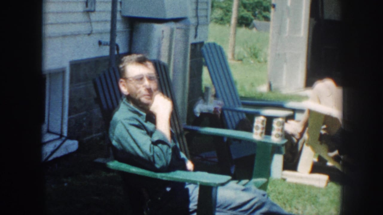 Man with glasses seated in a green outdoor chair beside a lawn and building.