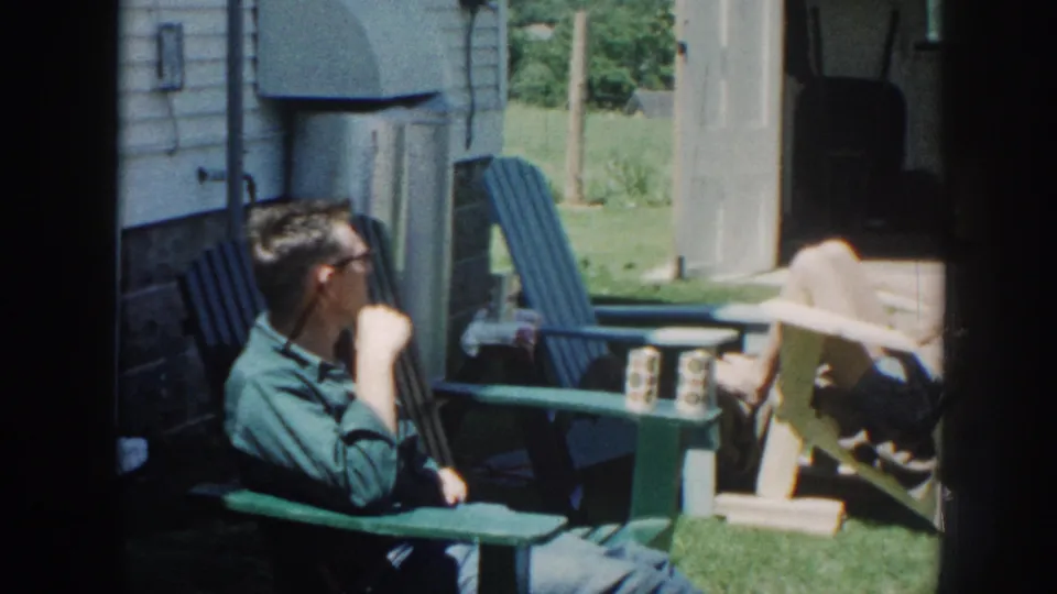 The man turns toward the reclining woman as they sit near a green table in a 1958 Nebraska backyard home-movie scene.