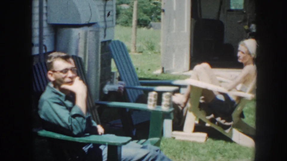 A man in glasses rests his hand near his face while a woman reclines nearby in the Nebraska backyard setting.