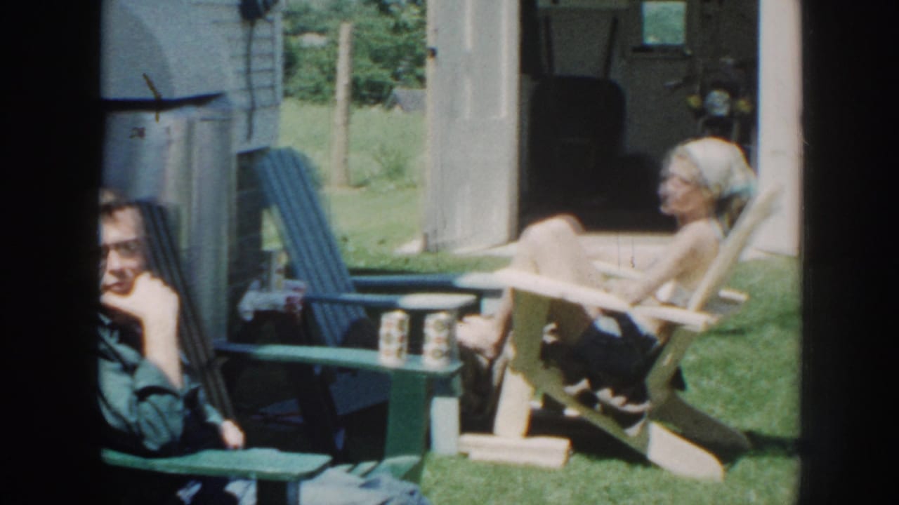 In 1958 Nebraska home-movie footage, two adults sit in Adirondack-style chairs near a garage and small table in a grassy yard.