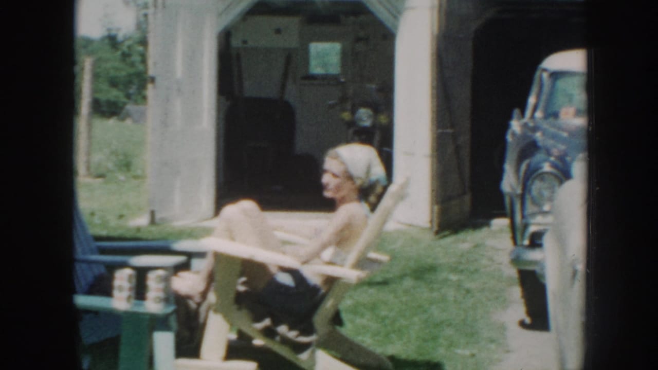 A woman in a head scarf sits in a pale outdoor chair near a parked vintage car.
