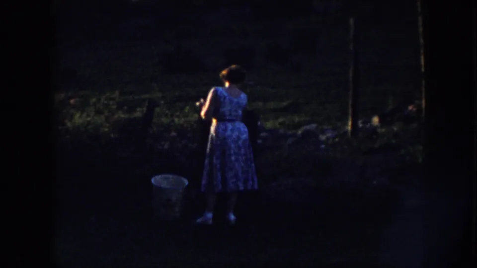 A woman stands in a dim outdoor garden area in the later part of the 1958 Nebraska home-movie clip, with a bucket on the ground nearby.