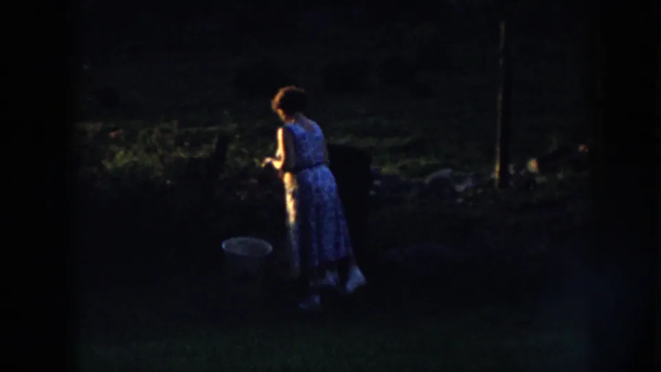 In 1958 Nebraska home-movie footage, a woman in a blue floral dress stands in a dim yard beside a large pale bucket.