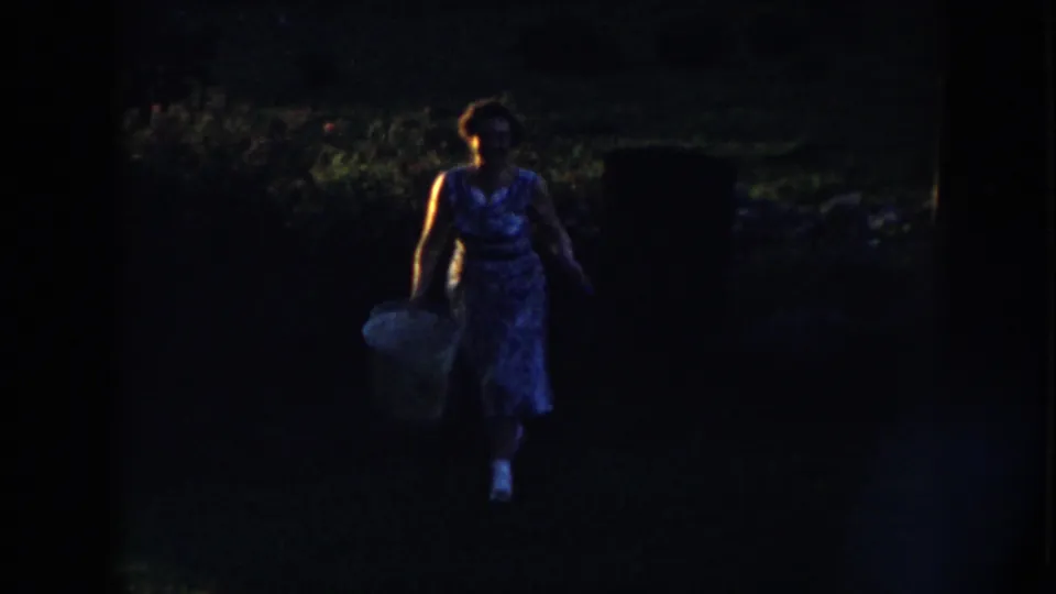 A woman in a blue floral dress walks through a dim Nebraska yard carrying a large pale bucket in 1958 home-movie footage.