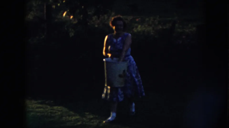 The woman approaches the camera with a large bucket, her floral dress and white shoes faintly visible in the dark 8mm frame.