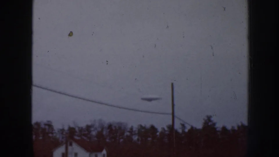 1958 Nebraska 8mm frame showing a dark disc-like object in an overcast sky above trees, a house roofline, a pole, and wire.