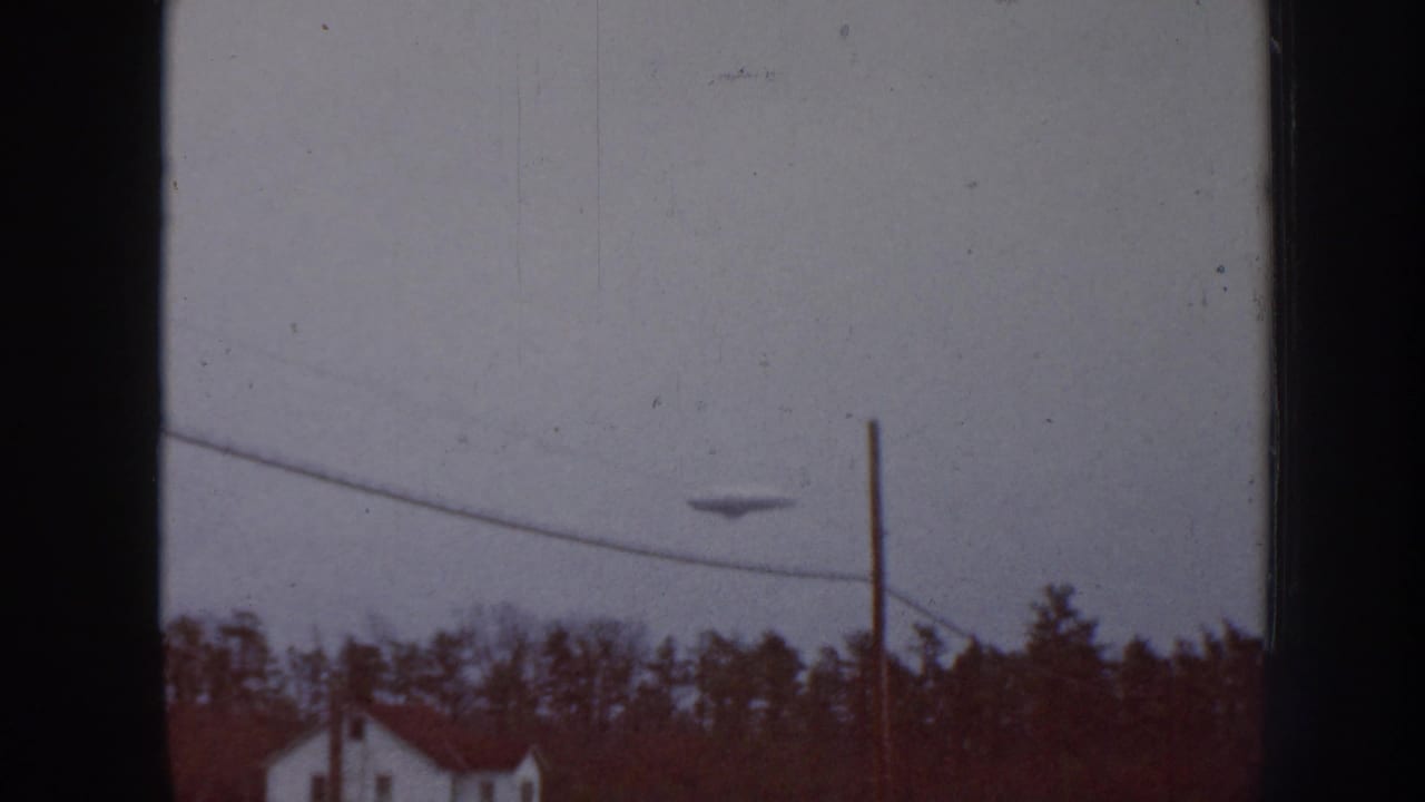 1958 Nebraska 8mm frame with a dark disc-like object near the center of the cloudy sky over a rural house, trees, pole, and wire.