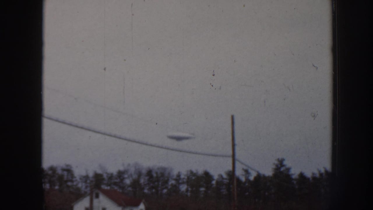 Disc-like object in a gray sky above utility wires, trees, and a distant house.