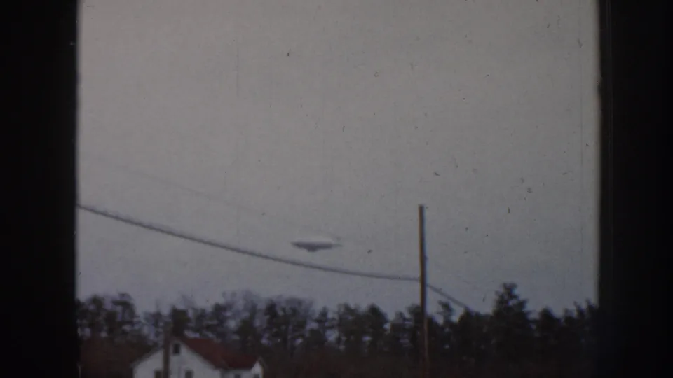 1958 Nebraska 8mm frame with a disc-like object in an overcast sky near a utility wire, pole, treeline, and distant house.