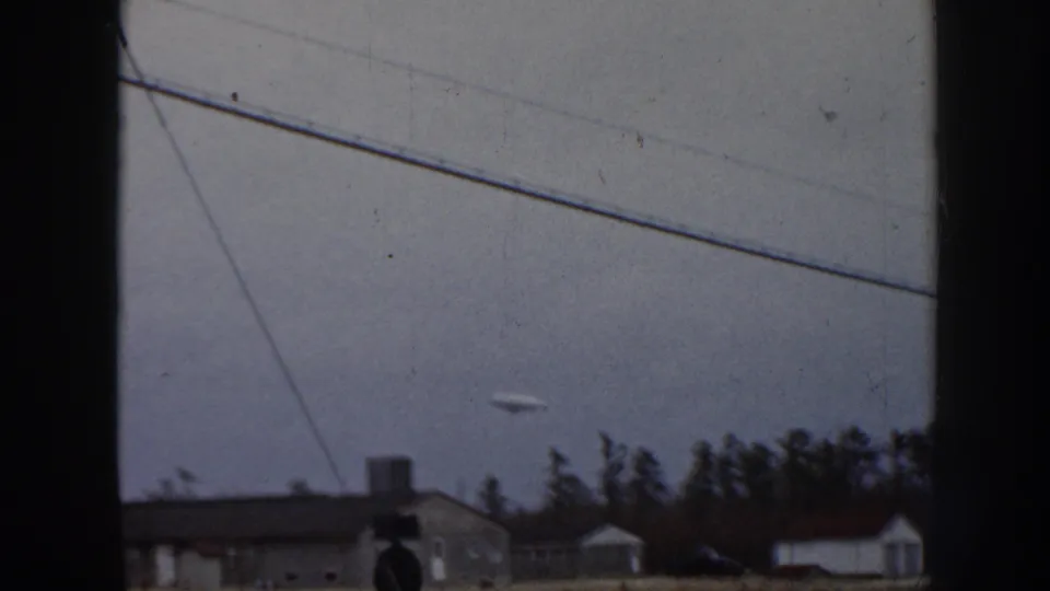 1958 Nebraska 8mm frame with the object lower in the sky near crossing utility lines, trees, rooftops, and small outbuildings.