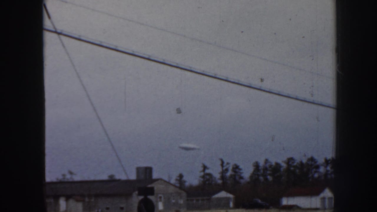 1958 Nebraska 8mm frame showing a small disc-shaped object in an overcast sky above low houses, tree silhouettes, diagonal utility lines, and dark window-frame edges.