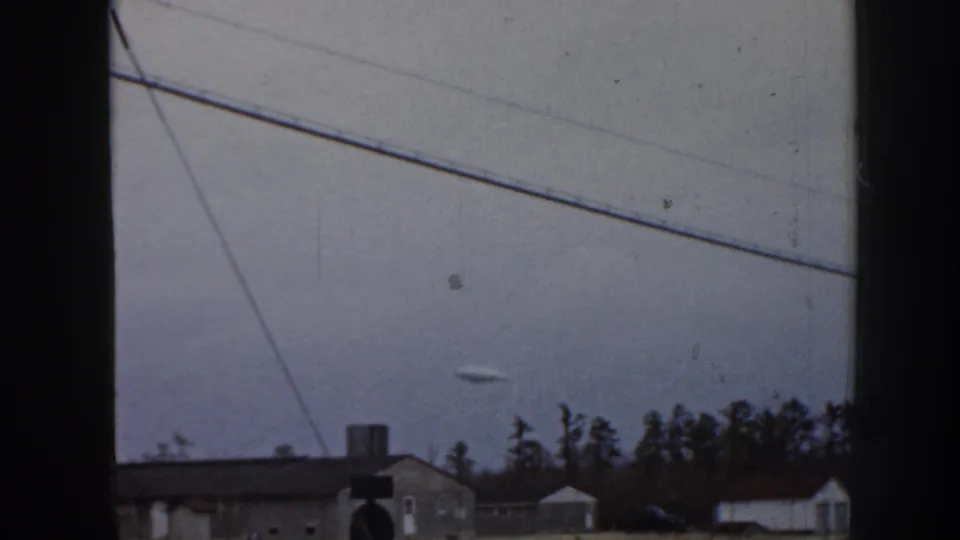 1958 Nebraska 8mm frame showing a small disc-shaped object in an overcast sky above low houses, tree silhouettes, diagonal utility lines, and dark window-frame edges.