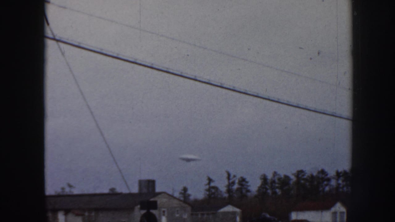 1958 Nebraska 8mm frame with a small unidentified disc-like object against a gray sky, crossed by utility lines above houses and trees.