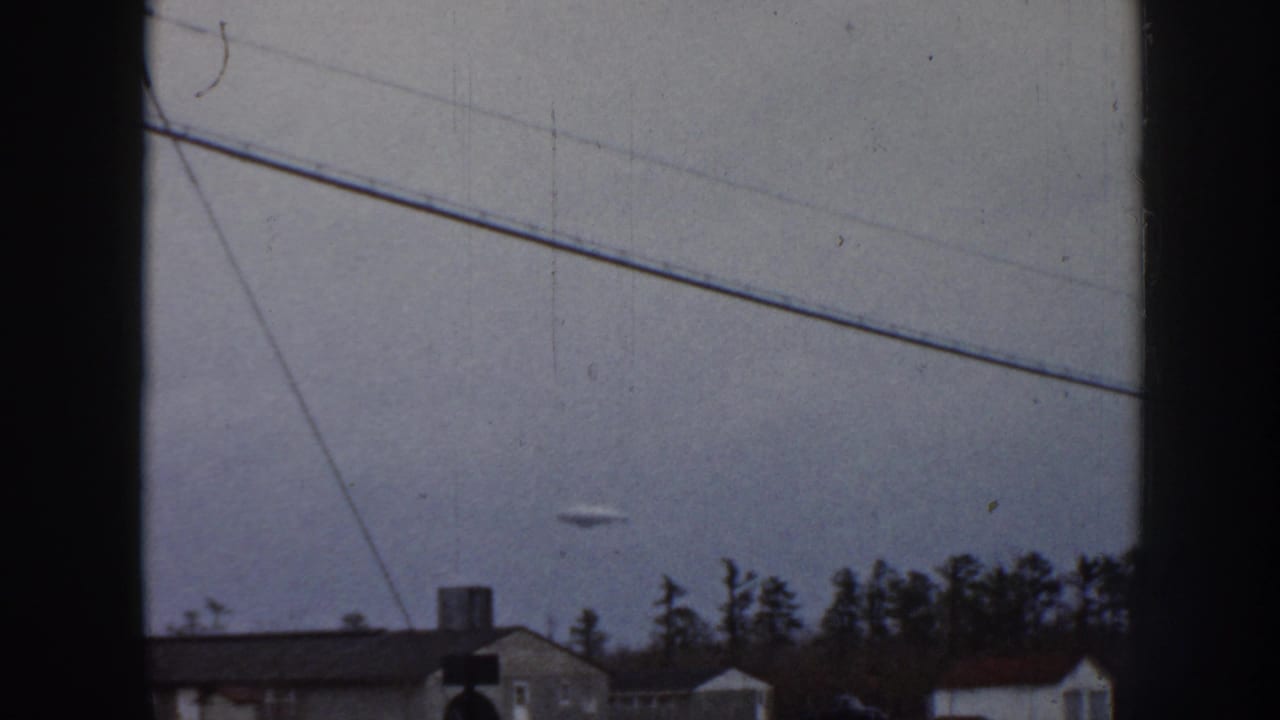 Small object in cloudy sky over a quiet neighborhood with trees and rooflines.