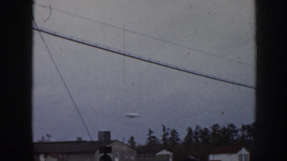 1958 Nebraska 8mm frame showing the disc-shaped object still visible in the cloudy sky, with power lines, house roofs, trees, and dark vertical framing in the foreground.