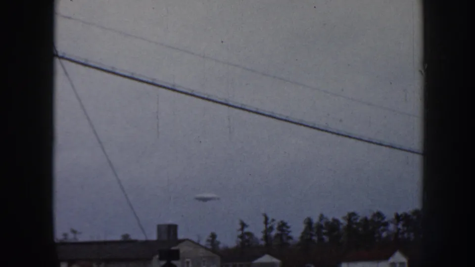 1958 Nebraska 8mm frame showing a snow-covered rural roadside, distant buildings, tree line, and pale winter sky.