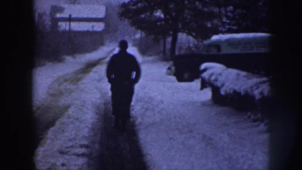The adult figure continues away from the camera on a snowy Nebraska road, with a parked vehicle and snow-covered roadside nearby.