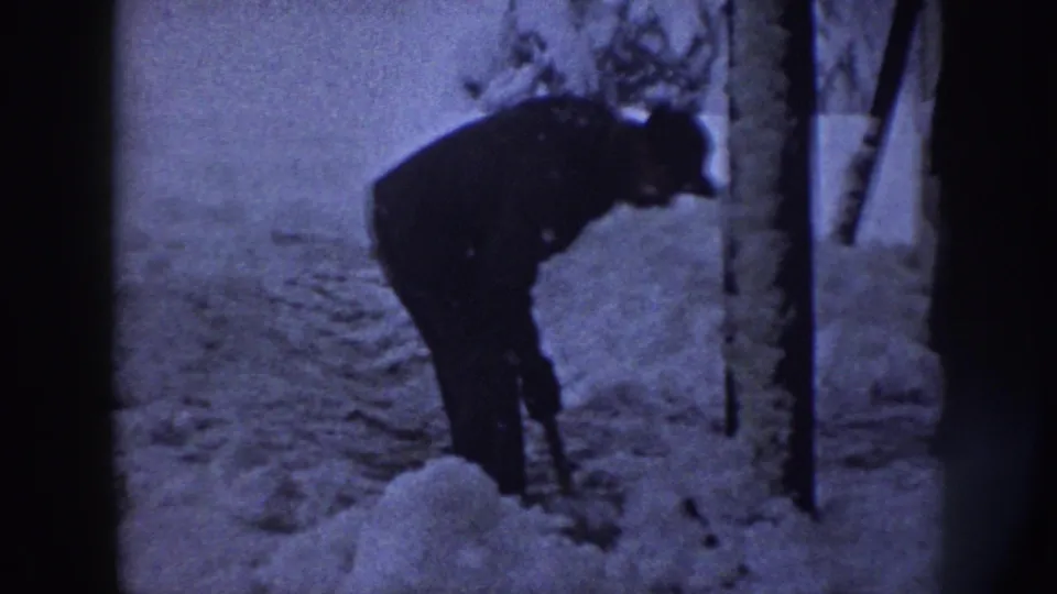 A man in dark winter clothing bends near roadside posts, appearing to shovel or clear snow in the rural Nebraska winter scene.