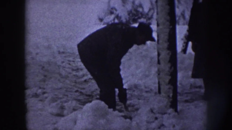 In 1958 Nebraska home-movie footage, an adult man in dark winter clothing bends to scoop snow near trees.