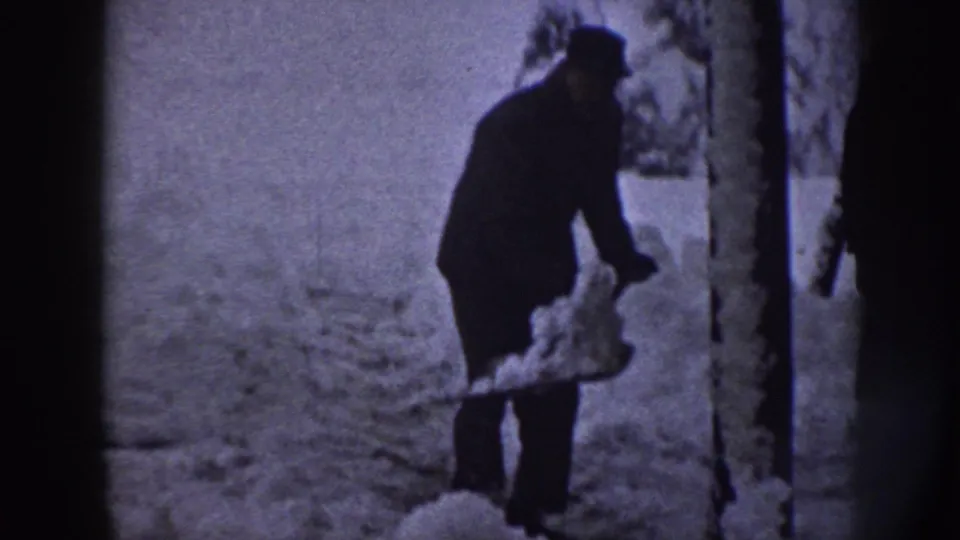 An adult man in a dark coat and cap lifts snow with a shovel among snow-covered trees in Nebraska, 1958.