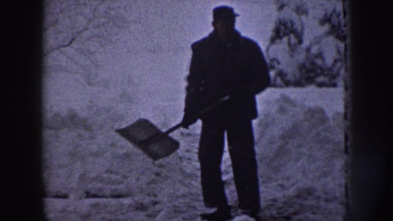 A man in dark winter clothing faces the camera with a shovel in a snowy wooded area, filmed on 8mm in Nebraska, 1958.