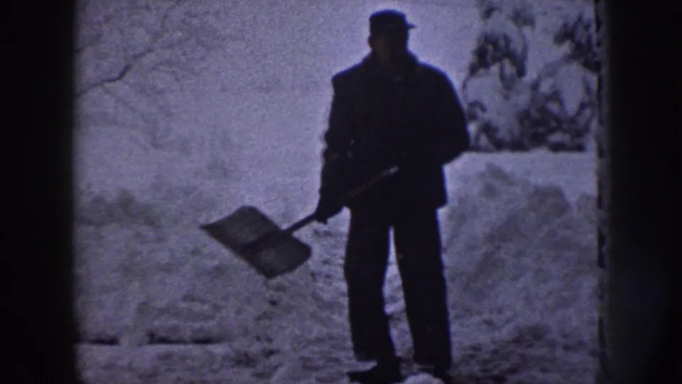 A man in dark winter clothing faces the camera with a shovel in a snowy wooded area, filmed on 8mm in Nebraska, 1958.