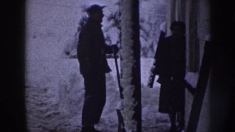 In 1958 Nebraska home-movie footage, two bundled figures stand in a snowy yard area, with one person holding a shovel.