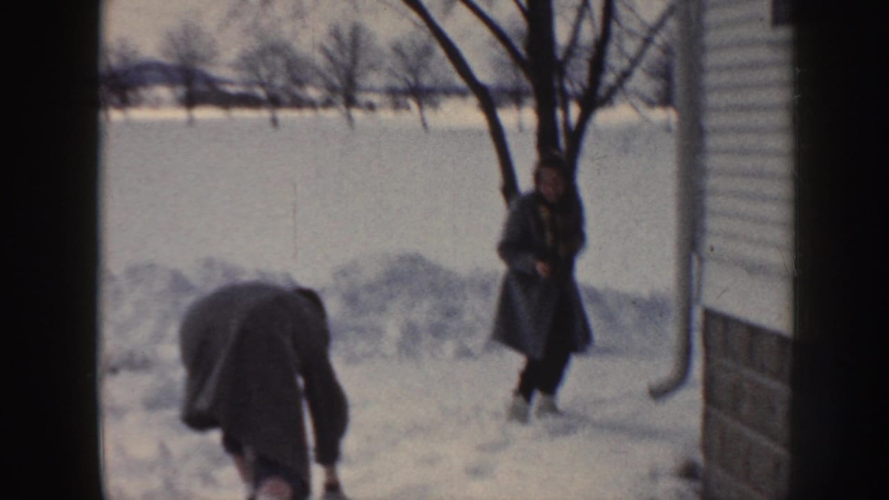 In a snowy Nebraska yard, one person bends down while another stands near a bare tree beside the exterior of a house.