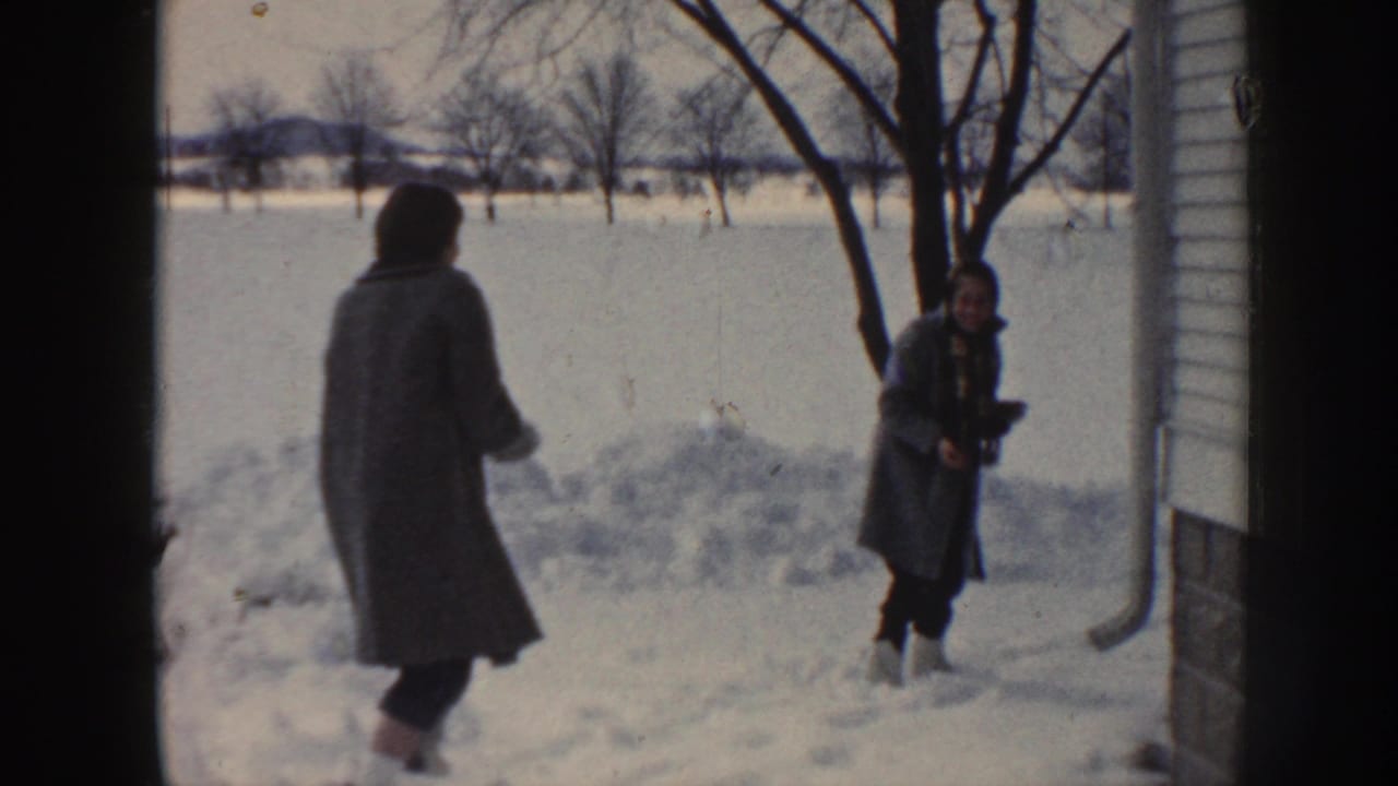 Two bundled people appear to play in a snow-covered Nebraska yard beside a house exterior and leafless tree.