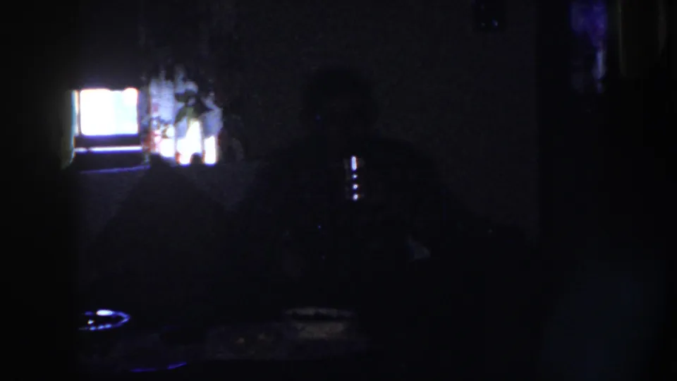 1958 Nebraska 8mm frame showing a dim private interior with a seated adult silhouette at a table beside a bright screen or window glow.