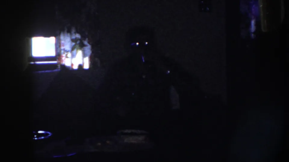 1958 Nebraska home-movie frame with an adult in deep shadow at a table, faint eye reflections, and a bright rectangular light behind him.