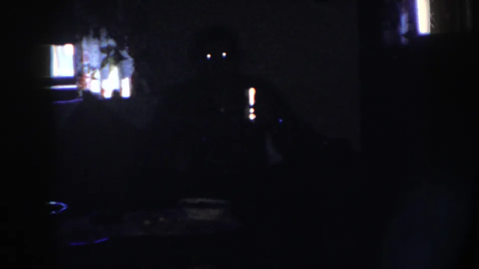 1958 Nebraska frame showing a dark private interior, a seated adult at a table, and a narrow bright reflection near the right side of the room.