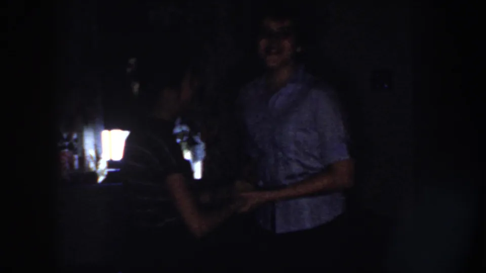 The couple dances close together in a dark Nebraska interior, with faces faintly visible in the 1958 8mm frame.